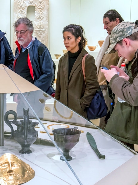 Visitors observing artifacts at the Archaeological Museum of Mycenae, including gold masks and ancient pottery.