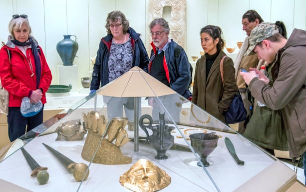 Visitors observing artifacts at the Archaeological Museum of Mycenae, including gold masks and ancient pottery.
