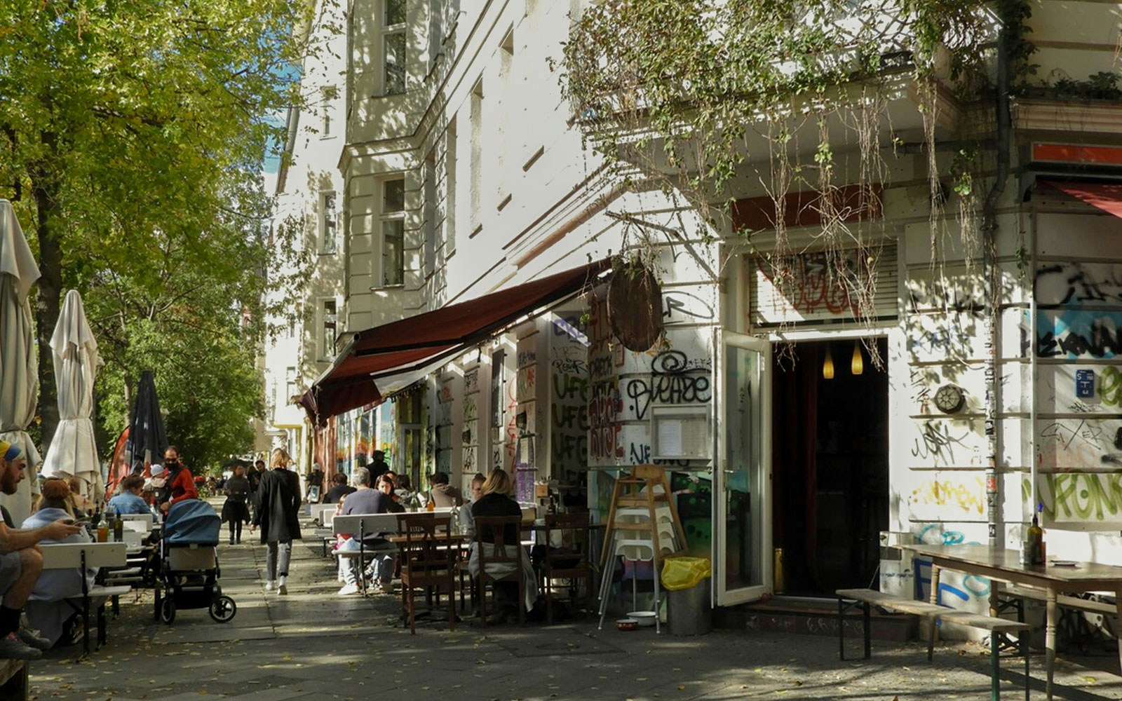 Eine belebte Berliner Straßenszene mit Café-Tischen im Freien, Graffiti an den Wänden und Menschen, die die Sonne genießen.