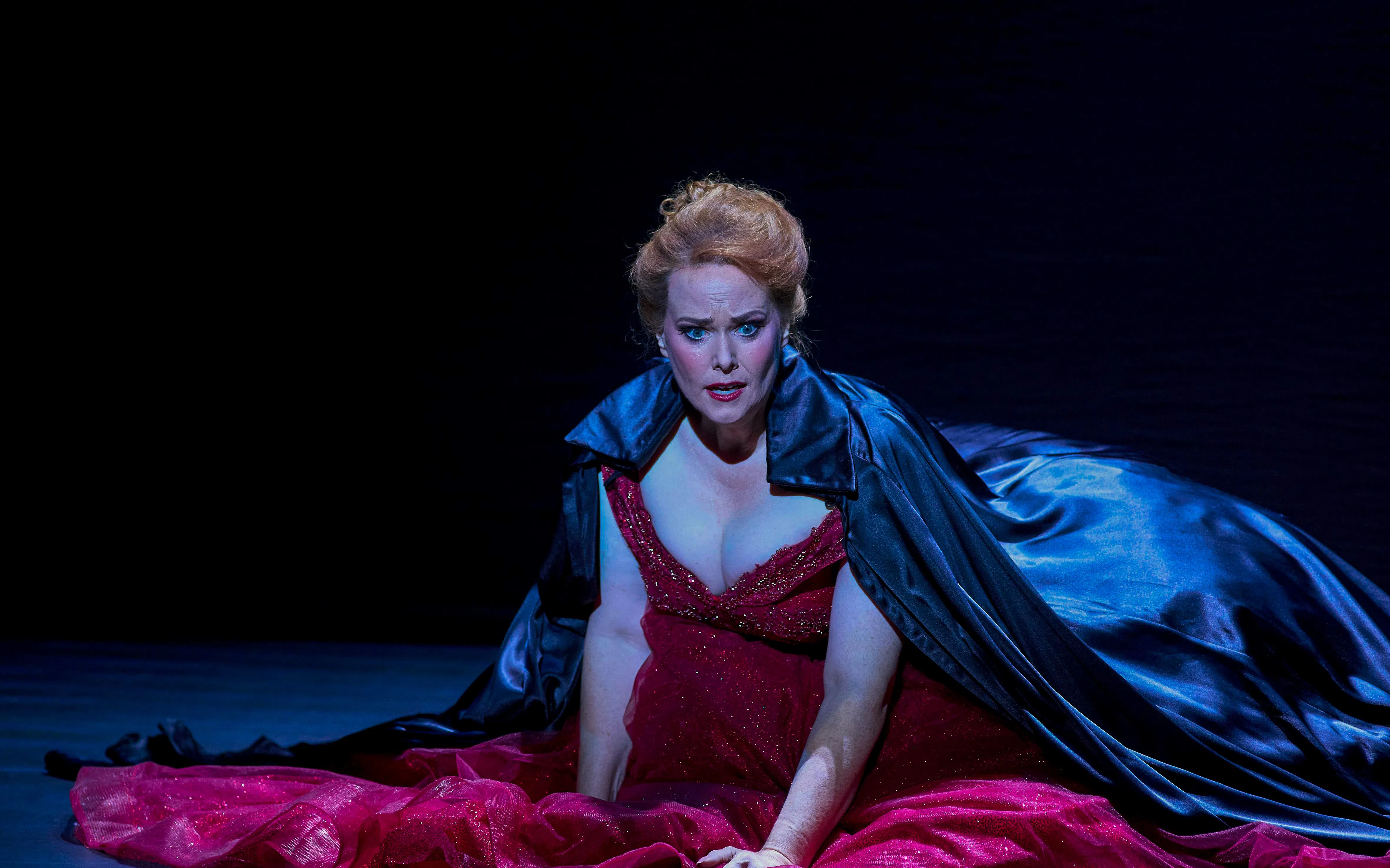 Performer in a dramatic scene from My Fair Lady, wearing a red gown and dark cape.