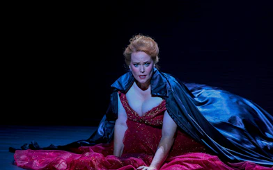 Performer in a dramatic scene from My Fair Lady, wearing a red gown and dark cape.