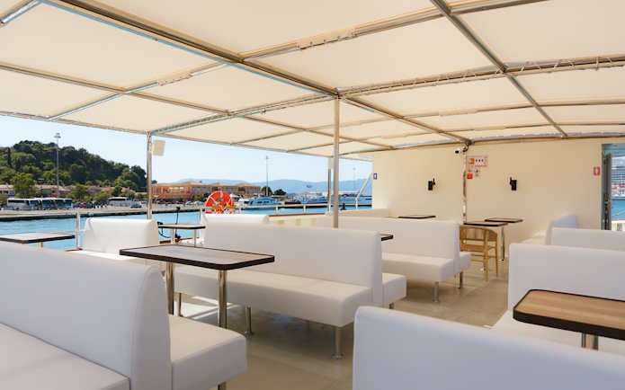 Corfu cruise boat interior with white seating and tables, docked at a scenic harbor.