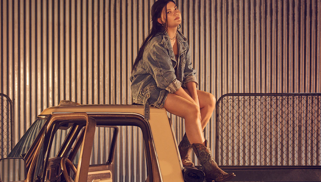 Person sitting on a truck roof in denim jacket and boots, with corrugated metal background.