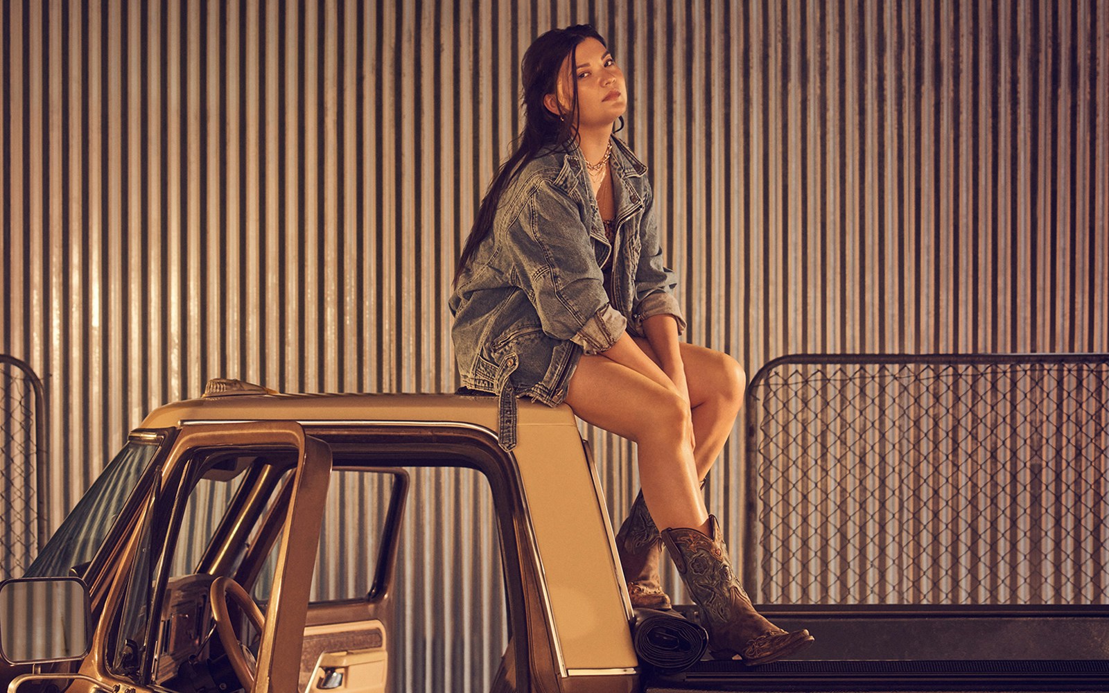 Person sitting on a truck roof in denim jacket and boots, with corrugated metal background.