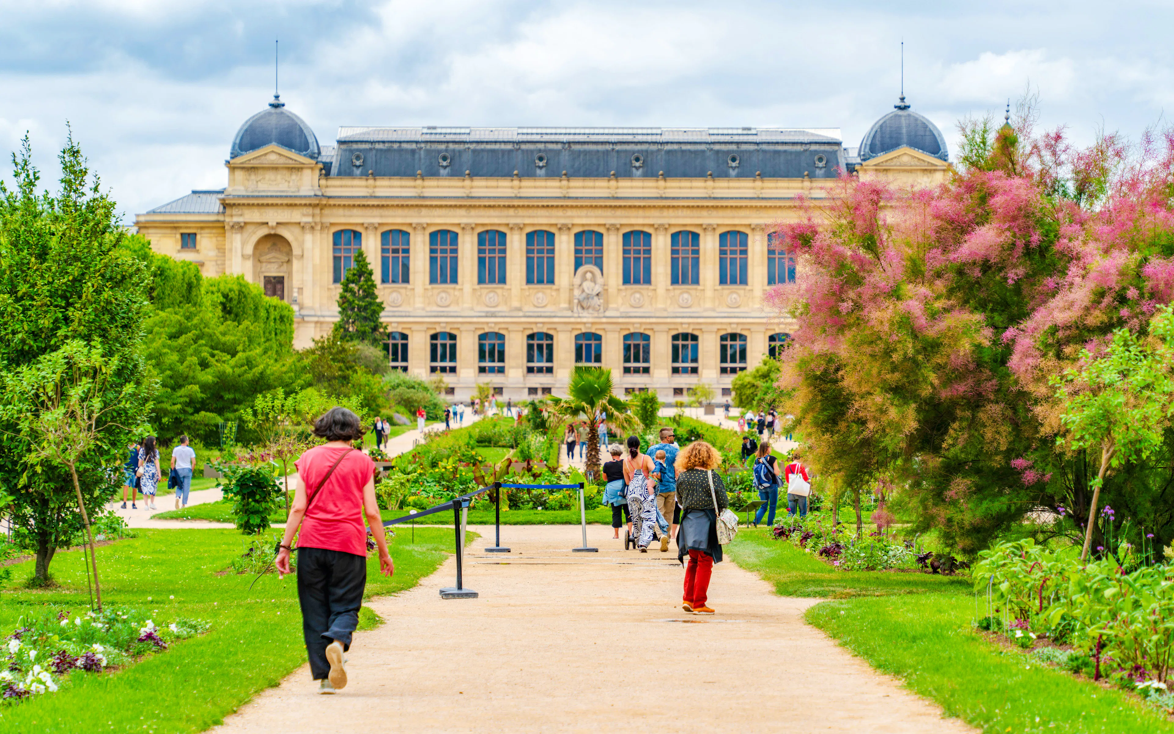 Visitors walking through Jardin des Plantes in Paris, France, with a historic building in the background.