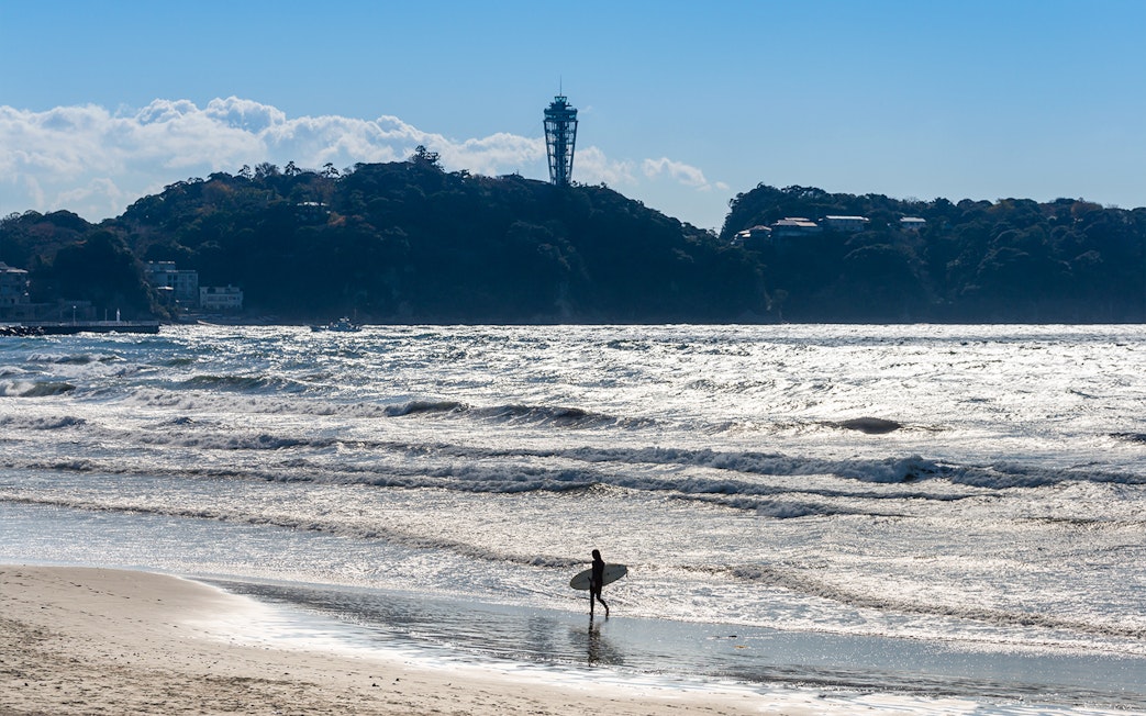 Surfer walking on Enoshima beach with Enoshima Sea Candle in the background.