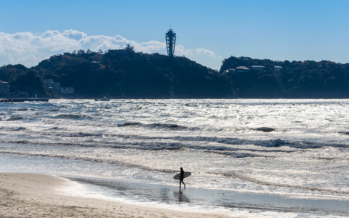 Surfer walking on Enoshima beach with Enoshima Sea Candle in the background.