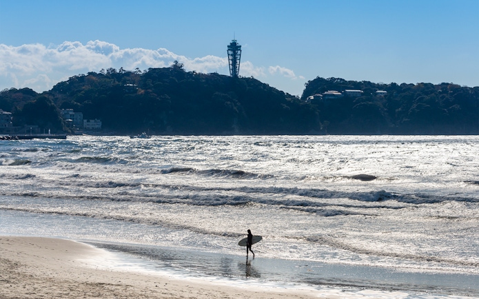 Surfer walking on Enoshima beach with Enoshima Sea Candle in the background.