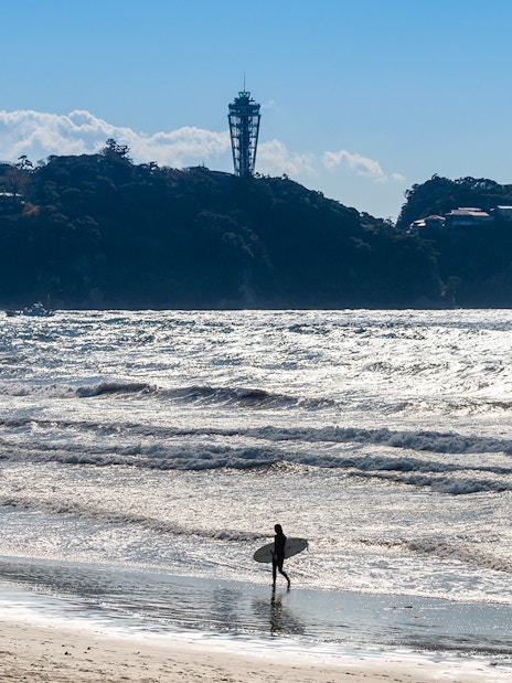 Surfer walking on Enoshima beach with Enoshima Sea Candle in the background.