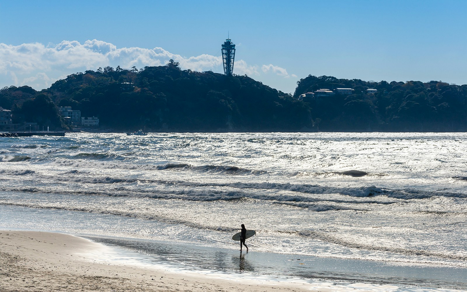Surfer walking on Enoshima beach with Enoshima Sea Candle in the background.