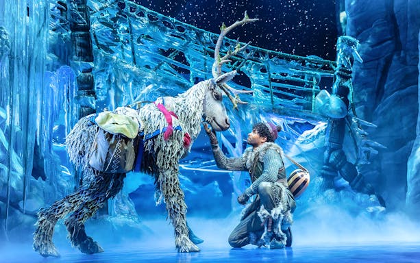 Man interacting with reindeer puppet on Disney FROZEN stage with icy backdrop.