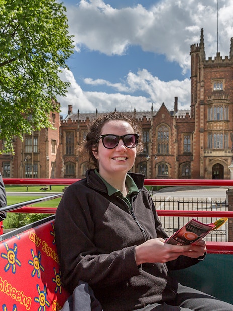 Hop-on hop-off bus near Queen's University Belfast with tourists taking photos.