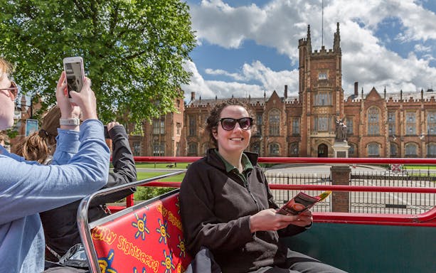 Hop-on hop-off bus near Queen's University Belfast with tourists taking photos.