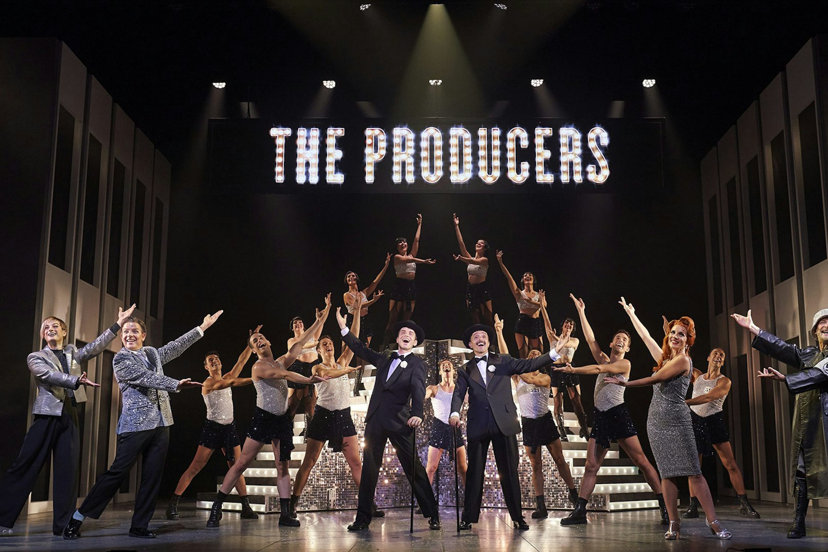 Cast performing a musical number from "The Producers" on stage with dancers and illuminated sign.
