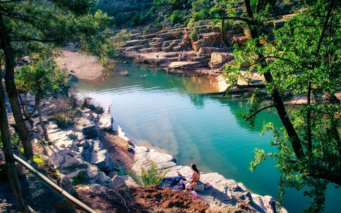 Rocky riverbank and clear water in Paradise Valley, Morocco, with a person sitting by the shore.