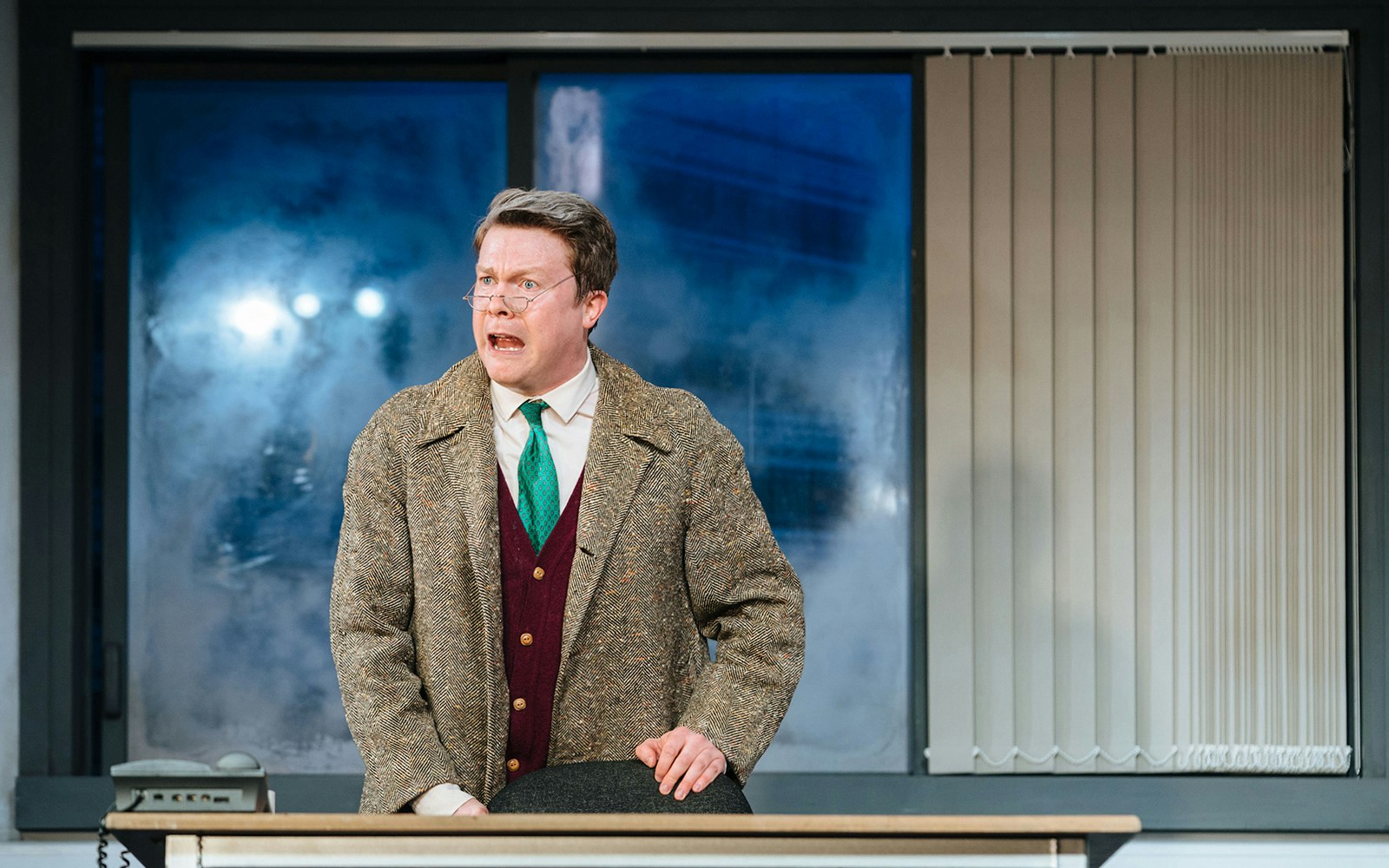 Man in a suit and overcoat speaking passionately at a desk, scene from "Accidental Death of an Anarchist.
