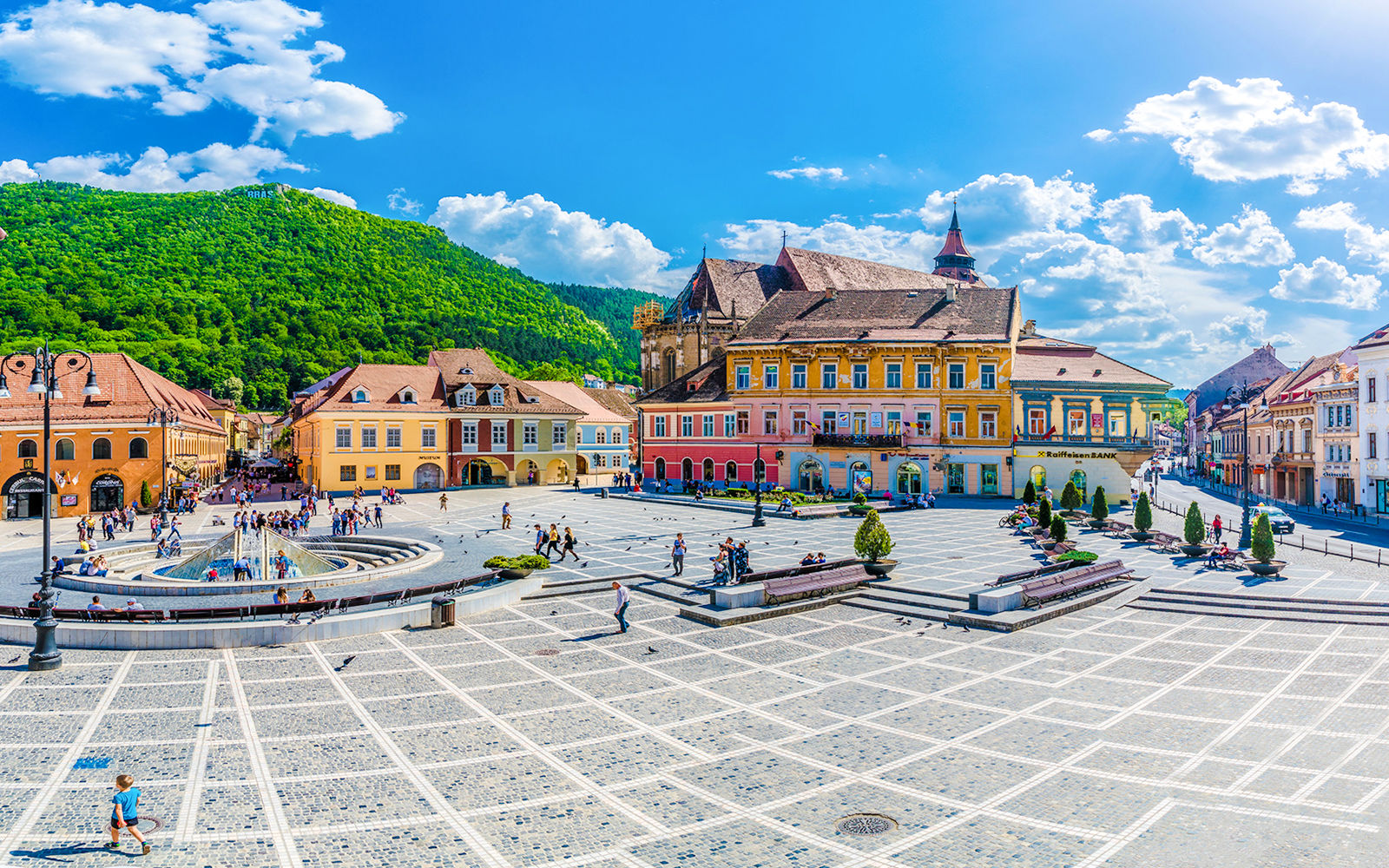 Brasov town centre with colorful historic buildings and a fountain, surrounded by green hills.
