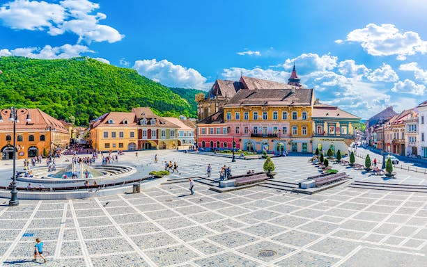 Brasov town centre with colorful historic buildings and a fountain, surrounded by green hills.