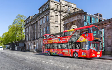 Red double-decker bus on Edinburgh Hop-On Hop-Off Tour near historic stone building.