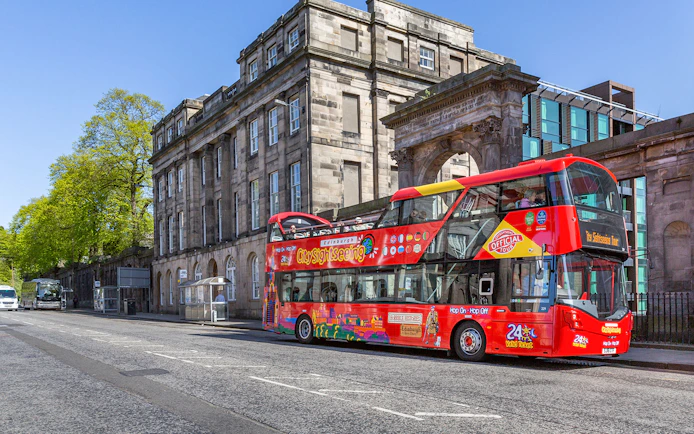 Red double-decker bus on Edinburgh Hop-On Hop-Off Tour near historic stone building.
