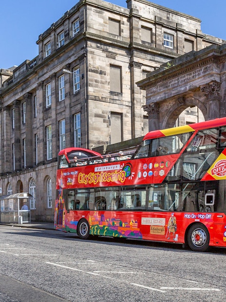 Red double-decker bus on Edinburgh Hop-On Hop-Off Tour near historic stone building.