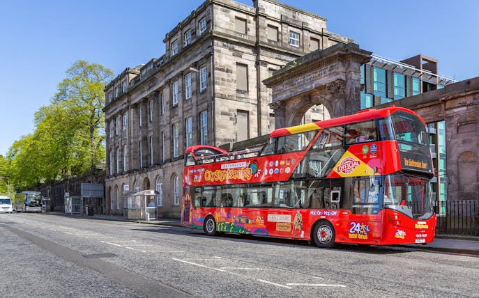 Red double-decker bus on Edinburgh Hop-On Hop-Off Tour near historic stone building.