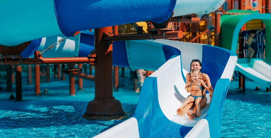 Mother and daughter sliding down a blue water slide into an outdoor pool.
