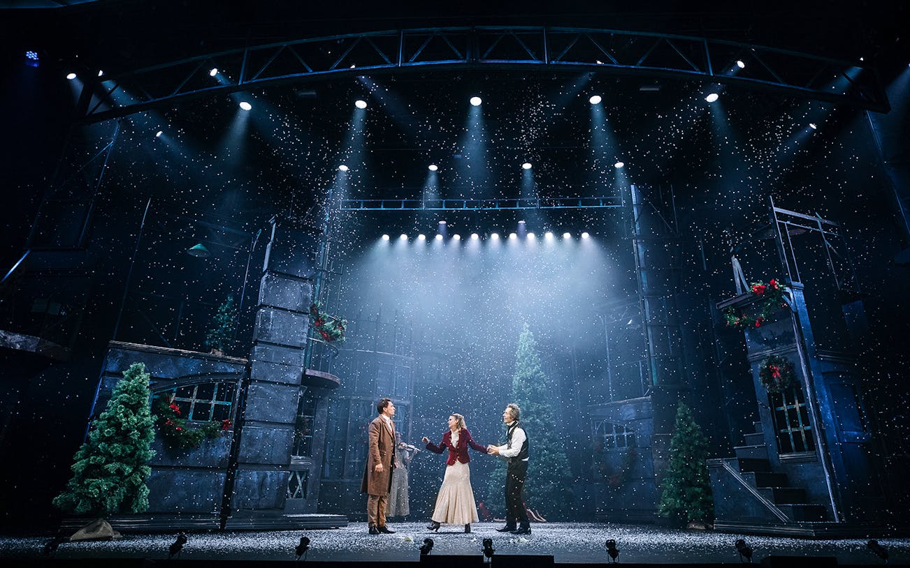 Stage performance of "A Christmas Carol" with actors in period costumes under snowfall.