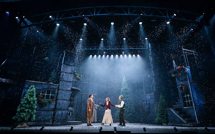 Stage performance of "A Christmas Carol" with actors in period costumes under snowfall.
