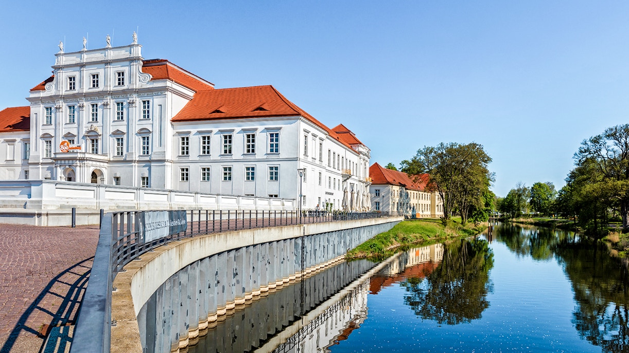 Schloss Oranienburg reflected in the Havel River, surrounded by trees.
