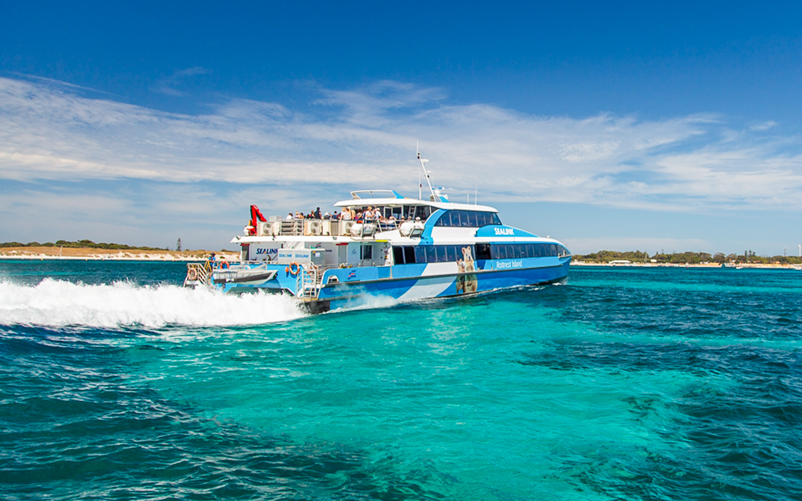 Eine Gruppe von Touristen genießt eine geführte Tour mit dem Bus auf Rottnest Island mit der Fähre im Hintergrund, die von Perth oder Fremantle zurückfährt.