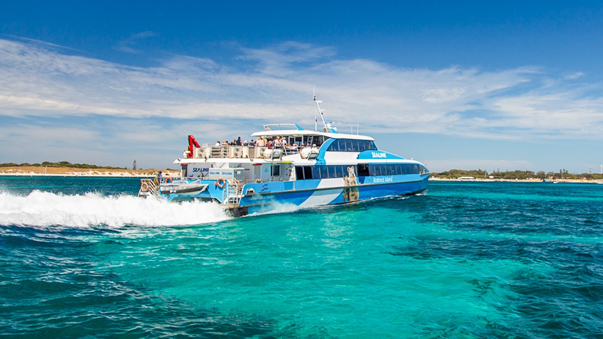 Group of tourists enjoying a guided bus tour on Rottnest Island with a return ferry visible in the background, departing from Perth or Fremantle