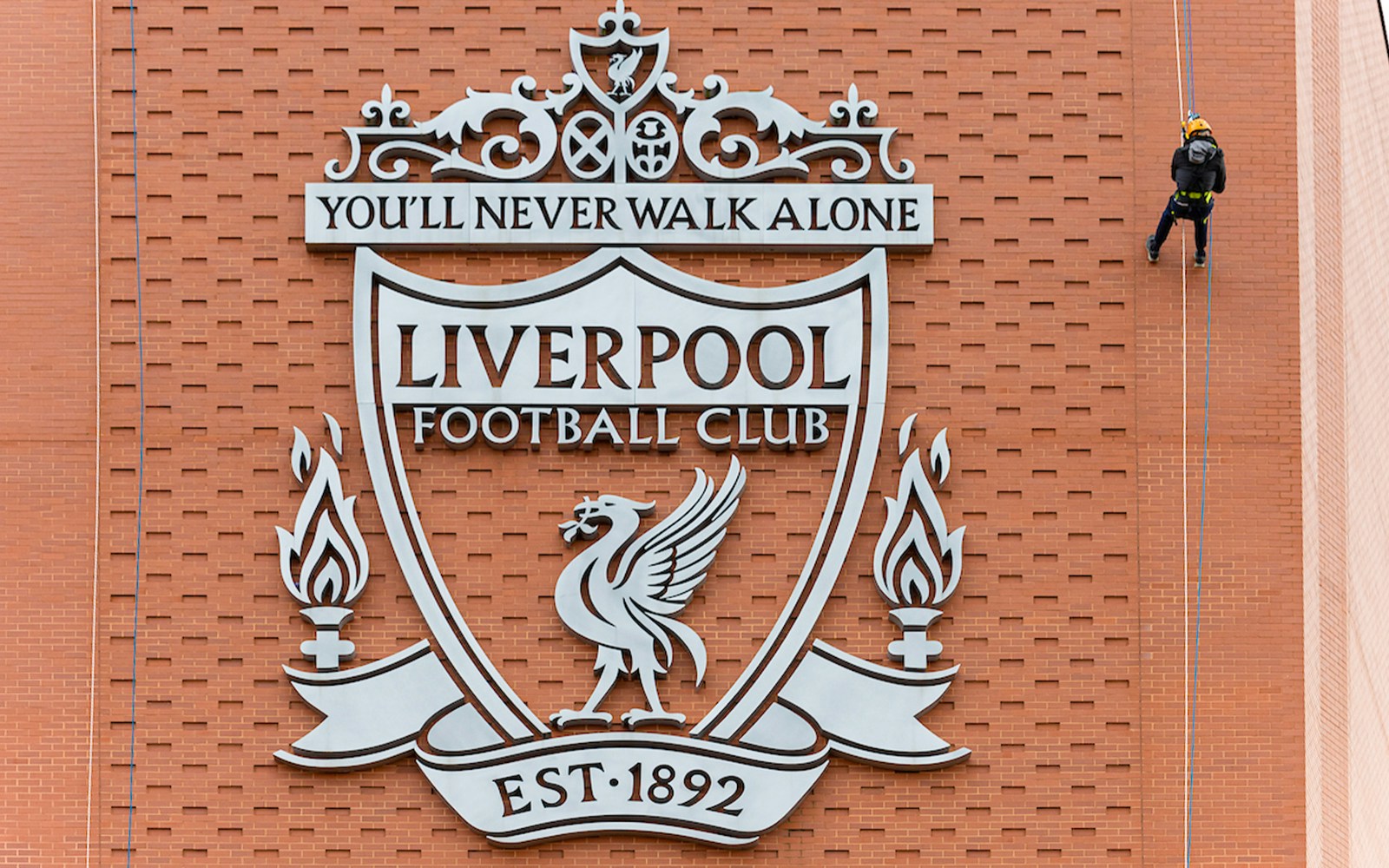 Abseiler descending Anfield Stadium wall beside Liverpool FC crest.