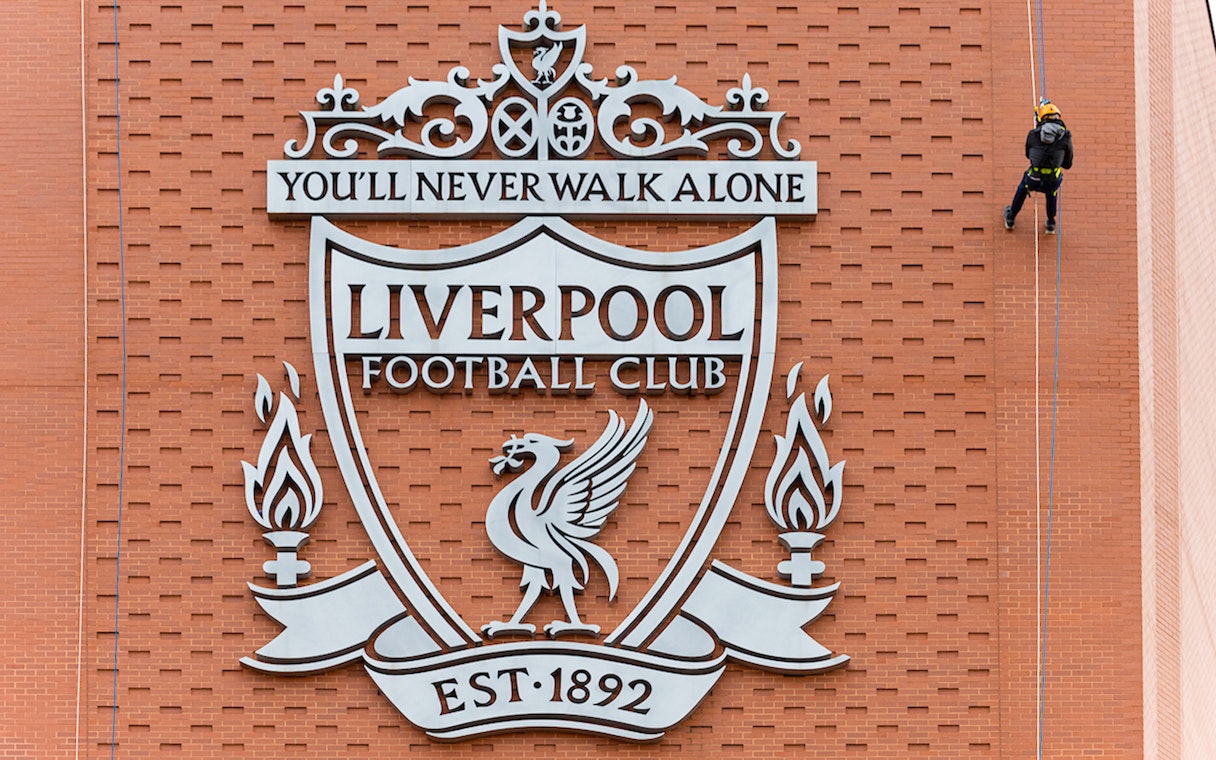 Abseiler descending Anfield Stadium wall beside Liverpool FC crest.