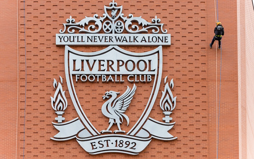 Abseiler descending Anfield Stadium wall beside Liverpool FC crest.