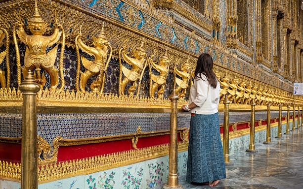 Woman admiring ornate golden garuda statues at Bangkok's Grand Palace.