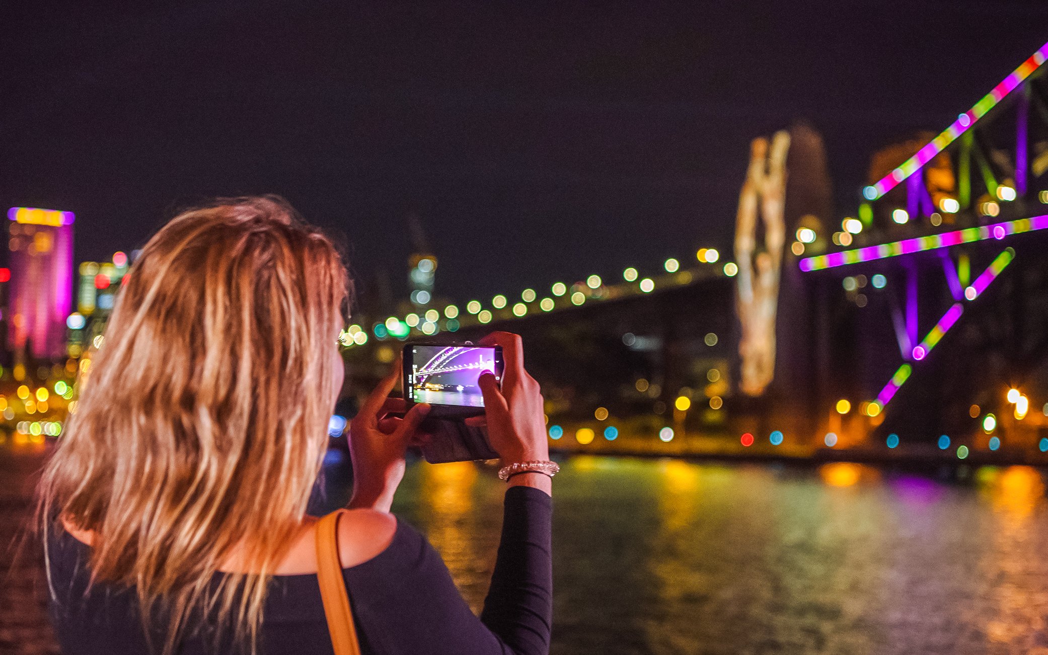 Person photographing Sydney Harbour Bridge illuminated during Vivid Sydney cruise.
