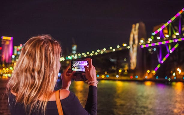 Person photographing Sydney Harbour Bridge illuminated during Vivid Sydney cruise.