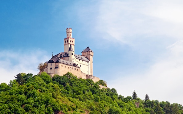 Marksburg Castle on a hilltop surrounded by lush greenery under a clear blue sky.
