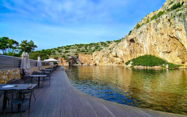 Vouliagmeni Lake in Attica, Greece with waterside dining area and rocky cliffs.