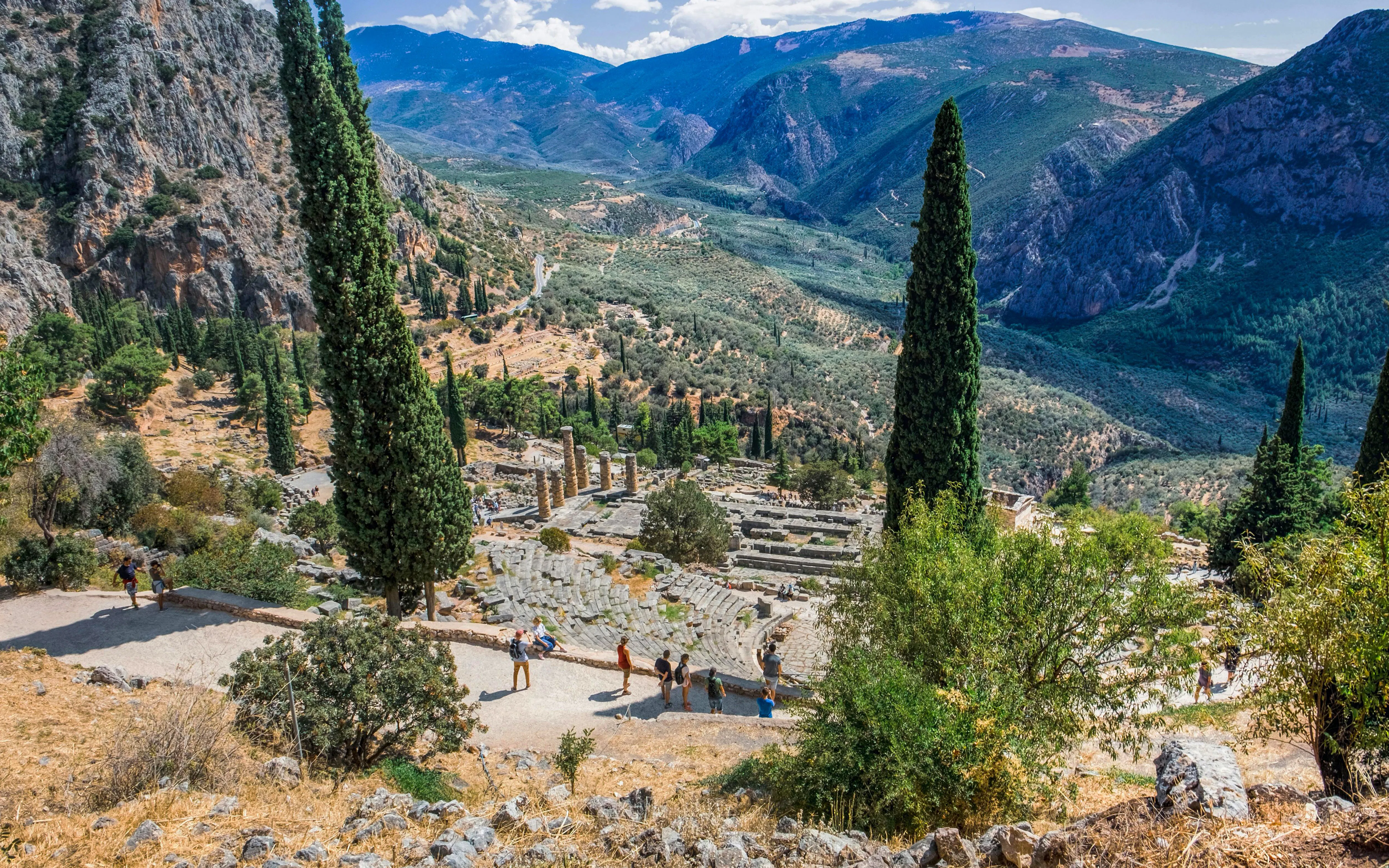 Tourists exploring the ancient ruins of the Temple of Apollo in Delphi, Greece, surrounded by mountains.
