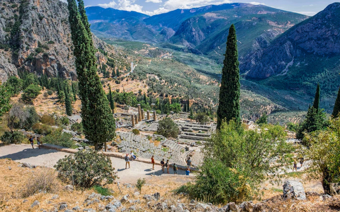 Tourists exploring the ancient ruins of the Temple of Apollo in Delphi, Greece, surrounded by mountains.