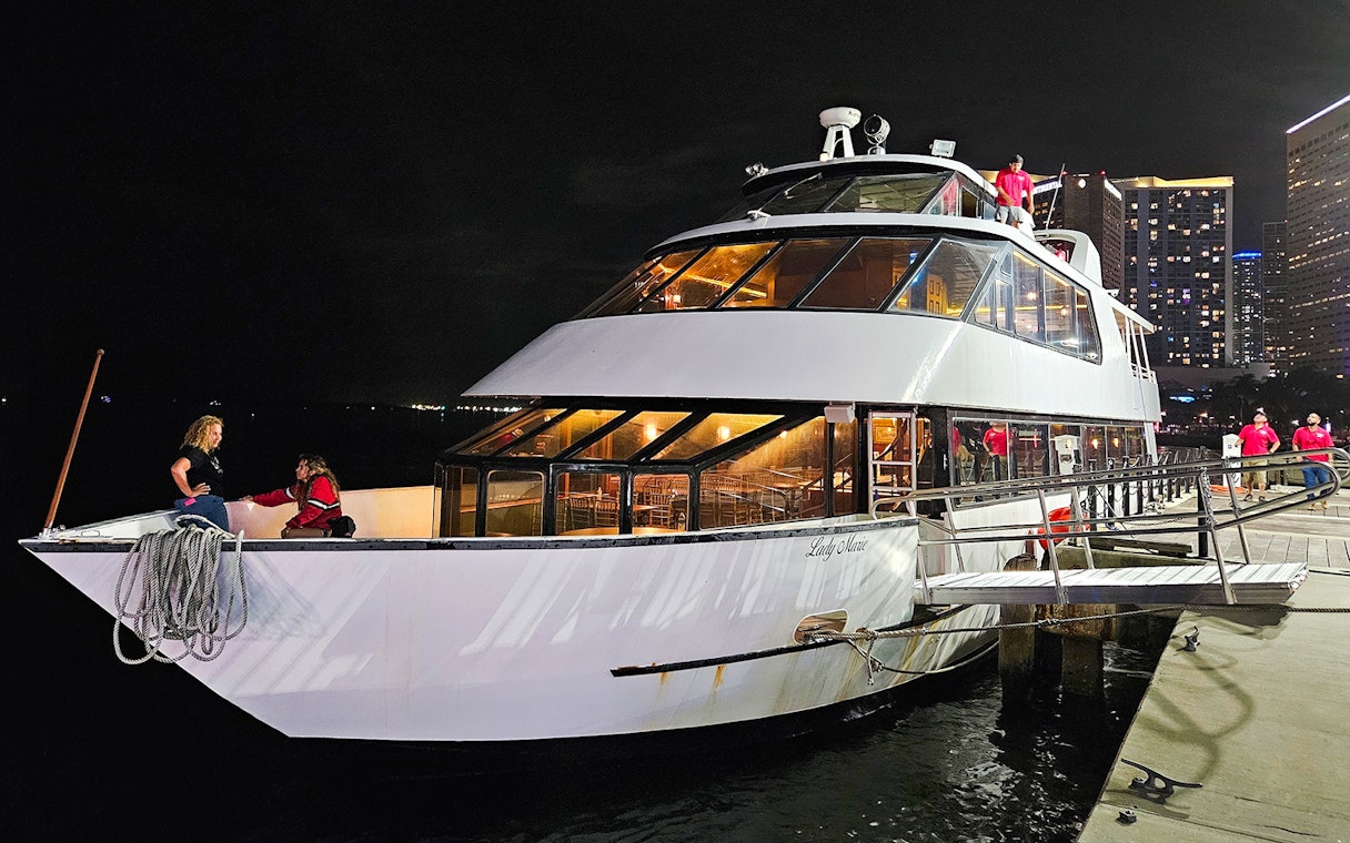 Party boat docked at night with people on deck, city skyline in background.