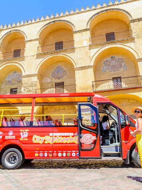Woman waiting outside hop-on hop-off mini bus in front of historic building in Córdoba, Spain.
