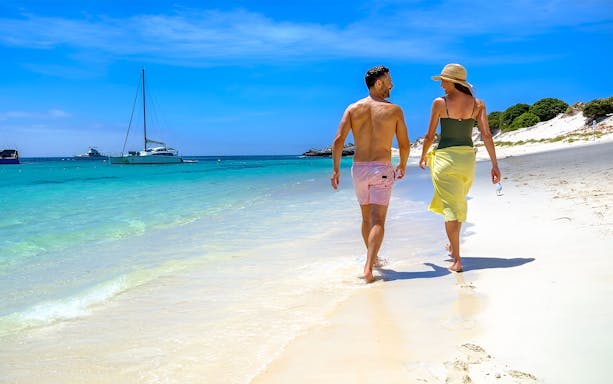 Couple walking on Rottnest Island beach with boats in the turquoise water.