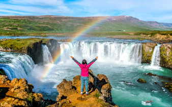 Person standing with arms raised in front of Godafoss waterfall, Iceland, with a rainbow overhead.