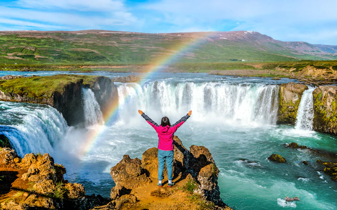 Person standing with arms raised in front of Godafoss waterfall, Iceland, with a rainbow overhead.