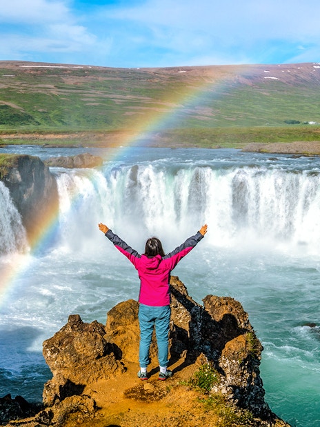 Person standing with arms raised in front of Godafoss waterfall, Iceland, with a rainbow overhead.