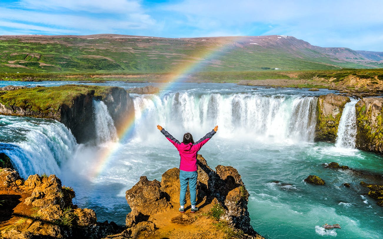 Person standing with arms raised in front of Godafoss waterfall, Iceland, with a rainbow overhead.