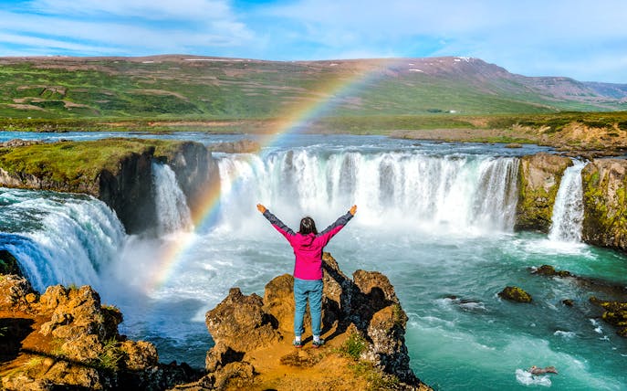 Person standing with arms raised in front of Godafoss waterfall, Iceland, with a rainbow overhead.